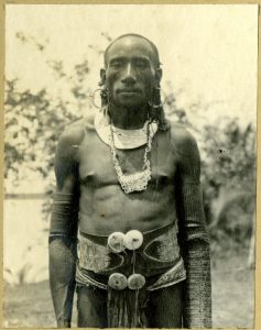 Fig.4 : Photographie (noir et blanc), d’un album ; portrait d’un homme en extérieur portant une ceinture d’écorce avec attaches de coquillages, collier de coquillages, boucles d’oreilles et ornements de bras : Papouasie-Nouvelle-Guinée. Epreuve à la gélatine argentique. Oc,A12.268, AN414768001 © The Trustees of the British Museum